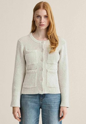 Kvinde med langt rødt hår iført en lys beige strikket cardigan med knapper og fire forlommer samt blå jeans.
