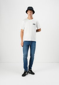 Young man wearing a black bucket hat, white Jack & Jones t-shirt, slim blue jeans, and black sneakers with white stripes stands against white background.