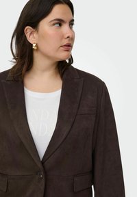 Pruun suede bleiser, millel on lõhikuga krae, ühe nööbi kinnitusega ja kaks esikotit, kombinatsioonis valge T-särgiga, millel on heledad tekstid.