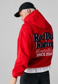Άτομο με πλατινέ ξανθά μαλλιά που φοράει μαύρο καπέλο και κόκκινο φούτερ με κουκούλα, με το κείμενο «Red Bull Racing Competing Since 2004» στην πλάτη, γκρι φόντο.