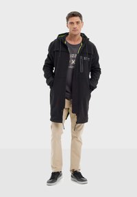 Homme portant un long manteau à capuche noir par-dessus un t-shirt sombre, un pantalon beige et des baskets noires, se tenant les mains dans les poches sur un fond uni.