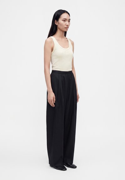 Femme aux longs cheveux foncés portant un haut beige sans manches et un pantalon noir à jambes larges, debout devant un fond gris clair uni.