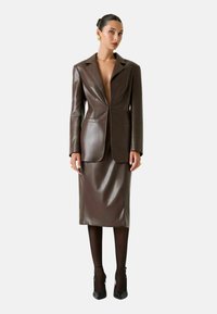 Blazer marrone in pelle con gonna a matita abbinata, caratterizzata da una profonda scollatura a V, chiusura con un bottone singolo e texture liscia. Completato da tacchi neri.