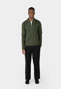 Mann som står iført mørkegrønn zip-up genser, svarte bukser og svarte sneakers med hvite såler mot en enkel lys bakgrunn.