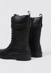 Black leather knee-high boots with a lace-up front, side zipper, and a strap with a horizontal buckle detail. Textured, smooth finish.