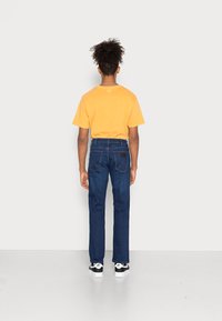 Camiseta de algodón amarilla y jeans de mezclilla azul oscuro. Los jeans cuentan con bolsillos traseros con detalles de costura. Zapatillas en blanco y negro completan el look.
