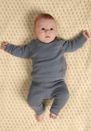 Baba texturált szürke kötött ruhában, krémszínű mintás takarón fekve, karjait kinyújtva, felfelé nézve.