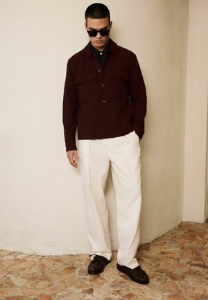Young man wearing dark brown buttoned jacket, white trousers, black shoes, and sunglasses standing against beige wall on stone floor.