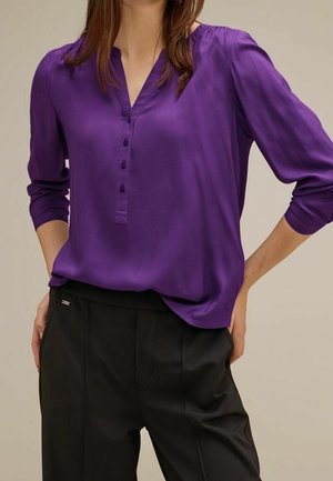 Blouse violette avec encolure en V, manches longues et fermeture à boutons. Texture lisse ressemblant à du satin, portée avec un pantalon noir sur mesure.