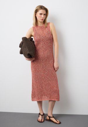 Donna che indossa un vestito midi rosa strutturato senza maniche e sandali marroni, che tiene in mano una grande borsa a maglia marrone scuro, in piedi contro un muro bianco.