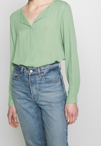 Blusa verde chiaro a maniche lunghe con scollo a V, abbinata a jeans azzurri a vita alta. Il tessuto sembra liscio e fluente.