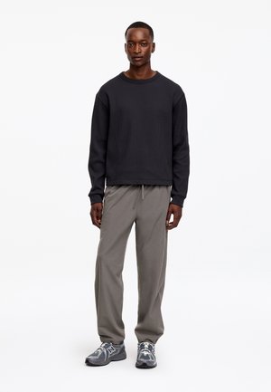 Homme debout portant un t-shirt noir à manches longues, un pantalon gris à cordon et des baskets New Balance grises sur un fond blanc uni.