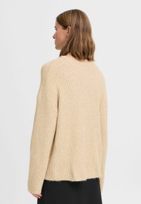 Beiger Rippstrickpullover mit lockerer Passform, langen Ärmeln, rundem Halsausschnitt und leicht verkürztem Saum. Weiche Textur und subtile Wärme.