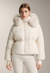 Woman wearing light beige puffer jacket with large fluffy fur collar and thumb hole cuffs, paired with beige leggings.