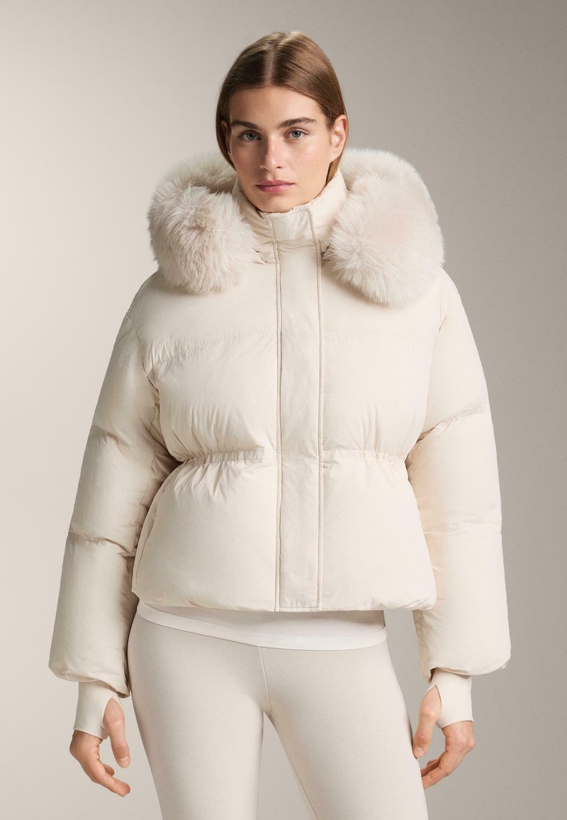 Woman wearing light beige puffer jacket with large fluffy fur collar and thumb hole cuffs, paired with beige leggings.