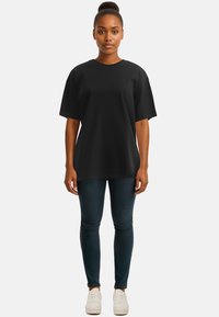 T-shirt nera a maniche corte con vestibilità rilassata e scollatura rotonda, abbinata a jeans scuri e sneakers bianche, in piedi davanti a uno sfondo neutro.
