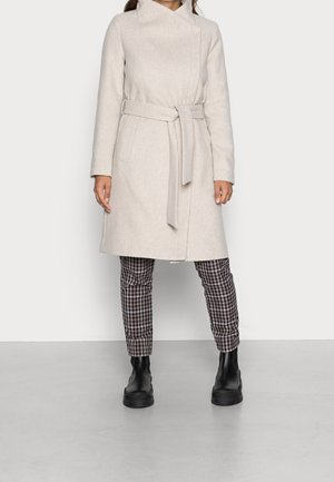 Persona con abrigo beige claro con cinturón, pantalones a cuadros y botines negros, de pie contra un fondo claro y liso.