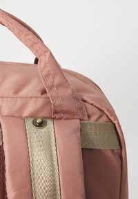 Sac à dos en tissu rose avec poignée supérieure, présentant une sangle texturée beige et des détails en laiton. Surface lisse avec bords cousus.