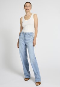 Žena stoji u svijetloplavim trapericama visokog struka, bijelom majicom bez rukava i smeđim sandalama s otvorenim prstima na jednostavnoj svijetloj pozadini.