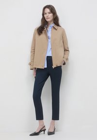 Frau in beigefarbenem Reißverschlussjacke über einem hellblauen Hemd, navyfarbenen Slim-Fit-Hosen und schwarzen Slingback-Pumps, die vor einem weißen Hintergrund steht.