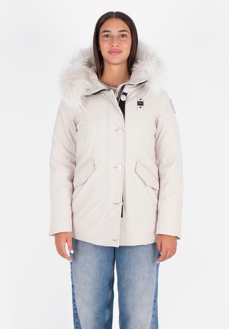 Chaqueta acolchada de un beige claro con capucha de pelaje, que incluye botones, bolsillos laterales y detalles en negro en contraste. Textura suave y corte relajado.