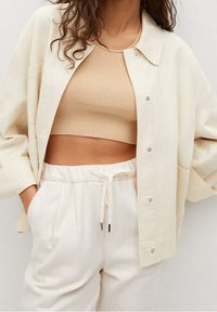 Jasna kremowa, oversize'owa koszula z zatrzaskami, noszona na beżowy, prążkowany top, w połączeniu z białymi spodniami z troczkami. Miękka faktura tkaniny.