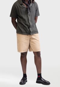 Olivgrünes Hemd mit kurzen Ärmeln und Knöpfen, mit strukturierten Streifen, kombiniert mit beigefarbenen Cordshorts. Schwarze Loafers und gemusterte Socken vervollständigen den Look.