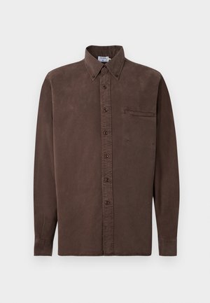 Άνδρας με κοντά μαλλιά που φοράει καφέ πουκάμισο με κουμπιά και γυρισμένα μανίκια, λευκό παντελόνι με φαρδιά μπατζάκια και μαύρα σανδάλια τύπου slides, στέκεται μπροστά σε απλό φόντο.