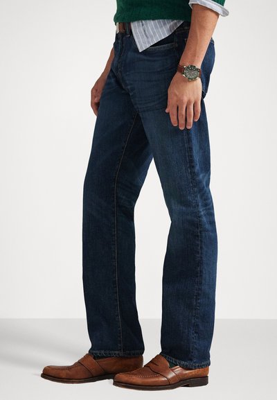 Dark blue denim jeans with a straight-leg cut, featuring contrasting stitching, and worn with brown leather loafers and a green knit sweater.