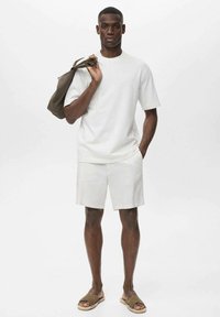 Un tricou alb cu maneci scurte si pantaloni scurti asortati, realizati dintr-un material neted. Modelul tine o geanta verde inchis pe umarul unui. Sandalele completeaza tinuta.