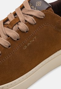 Brauner Wildledersneaker mit beigen Schnürsenkeln, mit einer strukturierten Oberfläche, einer weißen Zwischensohle und einem goldenen Logo auf der Zunge.