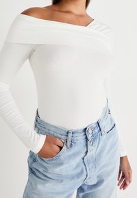 Žena v bílém dlouhém topu s odkrytými rameny, který má zastrčený do světlých modrých džín s vysokým pasem, s rukou v kapse.