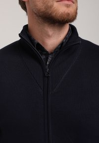 Jūrų laivyno zip-up megztinis su reljefine tekstūra, stovinčiu apykakle ir detaliomis petnešomis, dėvimas ant raštuoto marškinėlio.