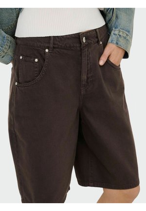 Kvinde iført mørkebrune løstsiddende denimshorts med forlommer, kombineret med en hvid top og en synlig denimjakkeærme.