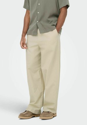 Homme portant un pantalon beige à jambes larges, des chaussures en suède marron et une chemise à manches courtes vert olive boutonnée, debout avec une main dans la poche.