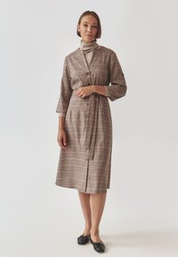 Vestito camicia a quadri marroni con maniche a metà, bottoni sulla parte frontale, cintura in vita e orlo fino al ginocchio, abbinato a ballerine nere.
