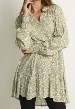 Woman wearing a light green floral long-sleeve dress with a V-neck and tiered skirt, posing with one hand near her face and the other on her waist.