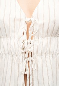 Κοντινό πλάνο ενός λευκού φορέματος με λεπτές καφέ κάθετες ρίγες και τρία δεμένα φιόγκους υφάσματος στη μέση μπροστινή άνοιγμα.