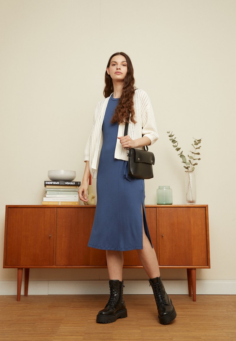 Vestido midi azul com fenda lateral, coberto por um cardigã branco tricotado. Modelo segura uma bolsa de ombro preta. Botas de combate pretas completam o visual.