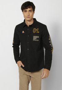 Camisa negra de manga larga con acentos gráficos amarillos, botones y un bolsillo en el pecho que presenta texto impreso y un logo triangular.