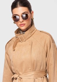 Femme portant des lunettes de soleil rondes marron, des boucles d'oreilles créoles dorées et un trench-coat beige ceinturé avec un col montant et des détails à boutons.