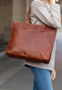 Bruine leren tote bag met twee handvatten, rechthoekige vorm, gladde textuur en subtiele stiksels, gedragen over de schouder.