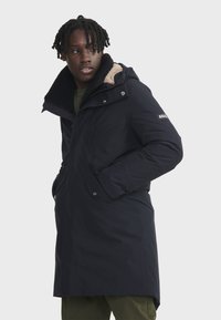 Manteau matelassé marine avec un col en fausse fourrure, fermetures éclair et à pression, et poches latérales. Présente un logo sur la manche et une coupe mi-longue.