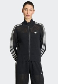 Crna pletena jakna na patentni zatvarač s mrežastom teksturom, visokim ovratnikom i bijelim trostrukim prugama duž rukava. Adidas logo na prsima.