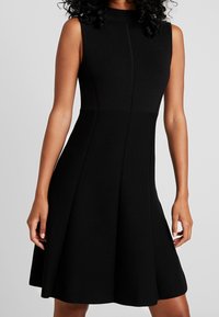 Robe noire ajustée sans manches avec jupe évasée portée par une personne ayant des cheveux bouclés sur fond neutre.