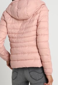 Veste matelassée rose avec un motif matelassé horizontal, capuche et poignets élastiques. Tissu doux et lisse avec une finition légèrement brillante.