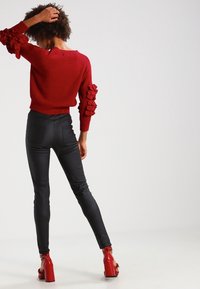 Punainen ribbineulottu neule röyhelöhihoilla, yhdistettynä mustiin istuviin housuihin ja punaisiin lohkopohjaisiin sandaaleihin. Tausta on vaalea ja yksinkertainen.