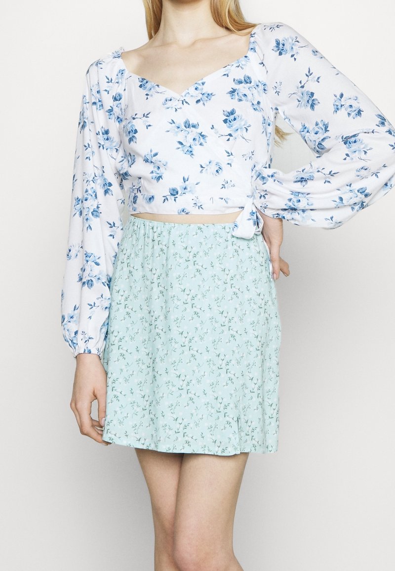 Femme portant un crop top blanc à manches longues avec un imprimé floral bleu et une jupe vert clair taille haute avec un petit motif floral.