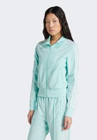 Giacca atletica azzurro chiaro con zip frontale, tre strisce bianche sulle maniche e logo sul petto, realizzata in materiale liscio e elastico.