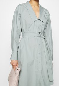 Svetle zelené šaty košilového střihu s širokým límcem, dlouhými rukávy, předními knoflíky a páskem uvázaným v pase; modelka drží strukturovanou tašku v odstínu broskve.